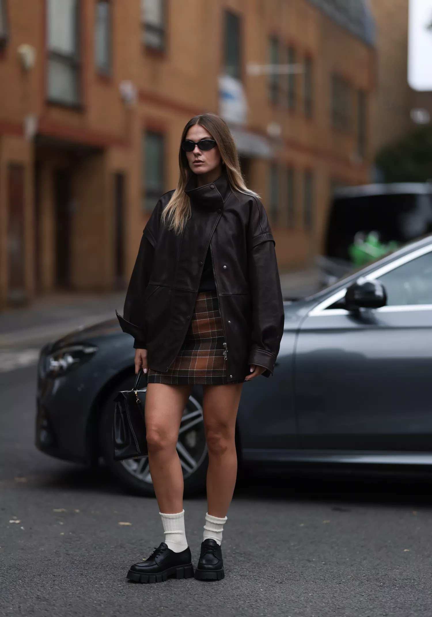 Alessa Winter is seen wearing black sunglasses from Oakley, a brown leather jacket from Gant, underneath a black top, a checked mini skirt in brown shades from Gant, a vintage Kelly bag from HermÃƒÂ¨s in black leather, white socks and black leather loafers from Marco Polo