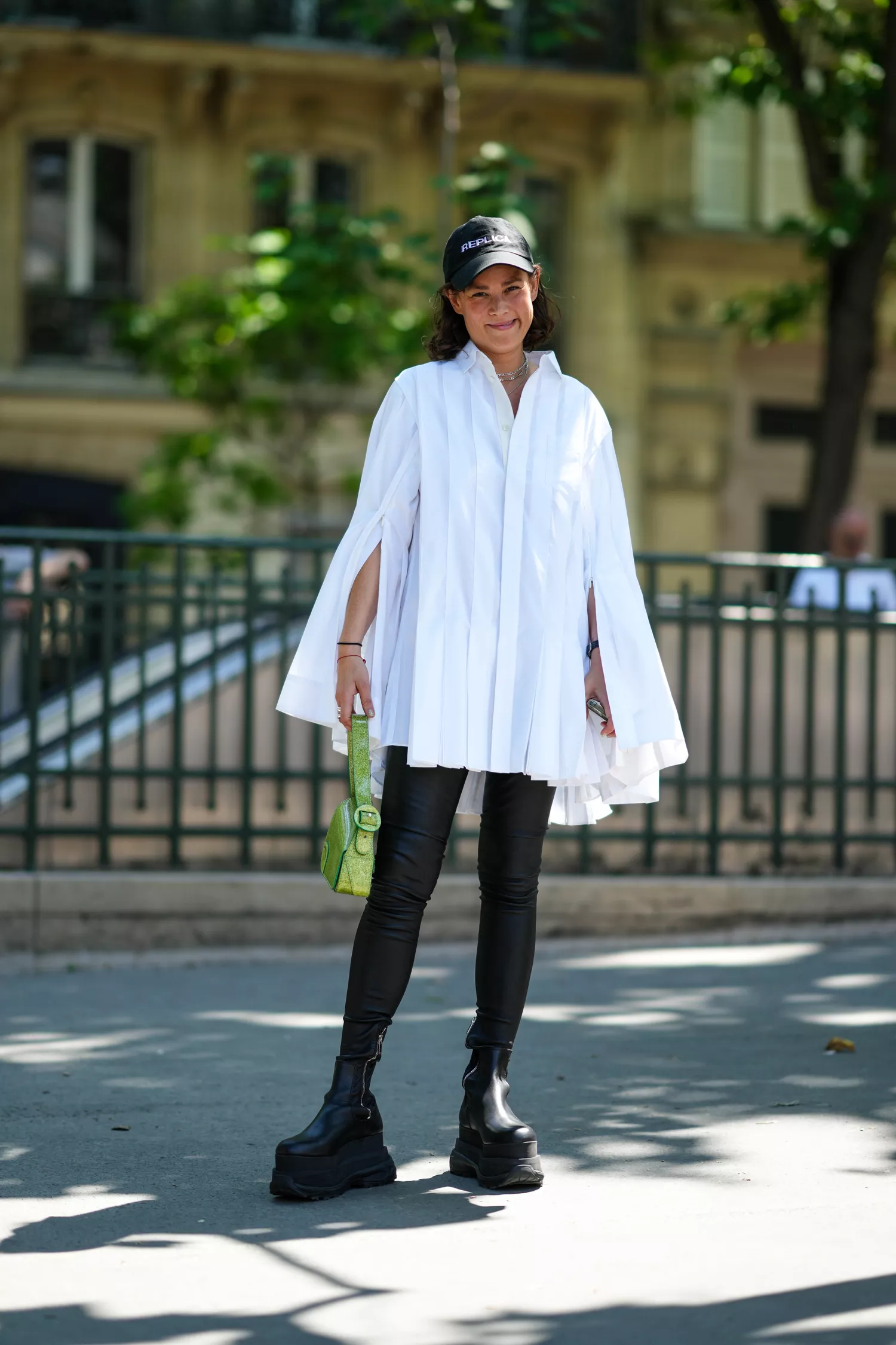 A guest wears a black with white Replica inscriptions pattern denim cap, silver large chains necklaces, a white pleated / accordion cloak shirt, black legging pants, a green glitter shiny leather handbag, black shiny leather platform soles ankle socks boots