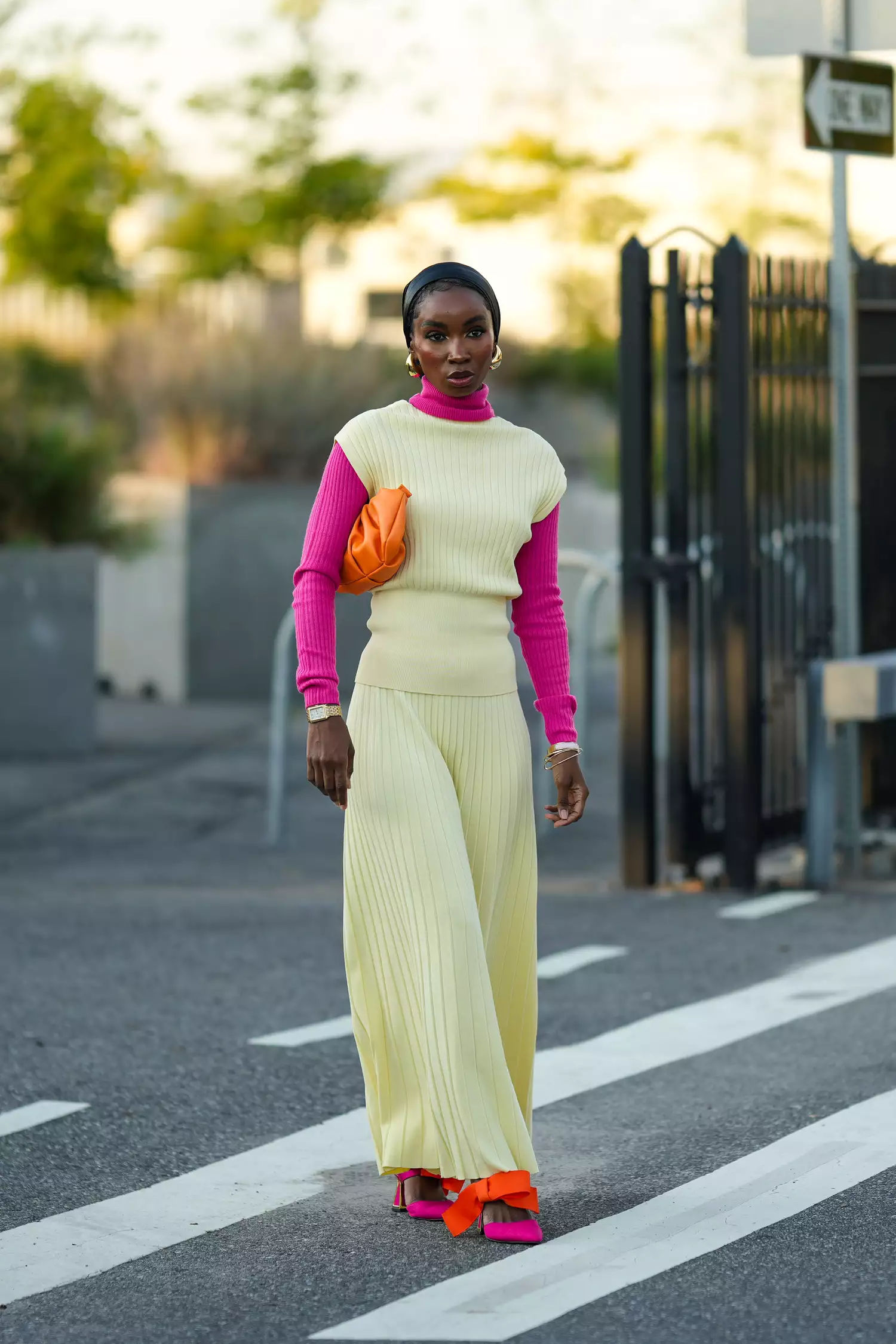 A guest wears a scarf over the head, a neon pink turtleneck top, a yellow pleated sleeveless long dress, pink and orange pointed shoes with attached bow ties