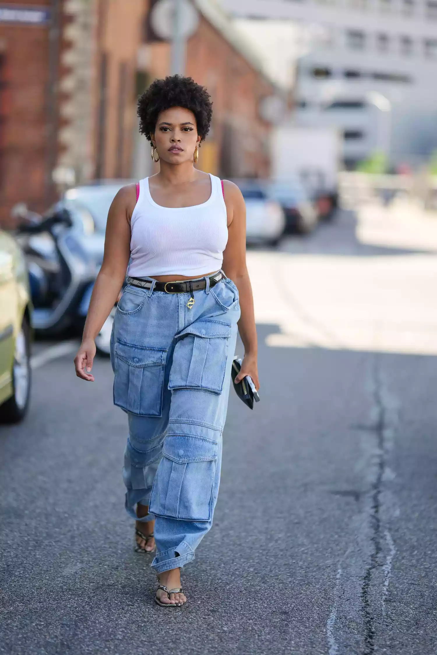 A guest wears a white tank top, a belt, blue denim cargo pants with large pockets, sandals, earrings