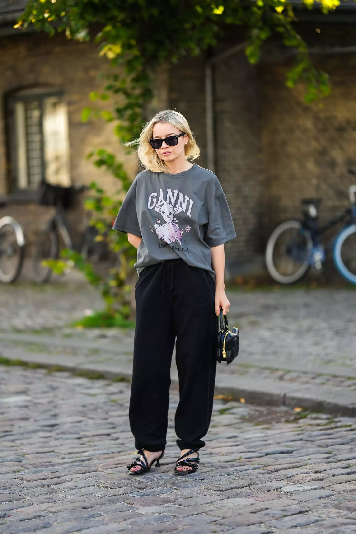 model wears black sunglasses, gold micro earrings, a pale gray with embroidered white and pale pink pattern t-shirt from Ganni, black large pants, a black shiny leather zipper handbag, black shiny leather asymmetric heels shoes