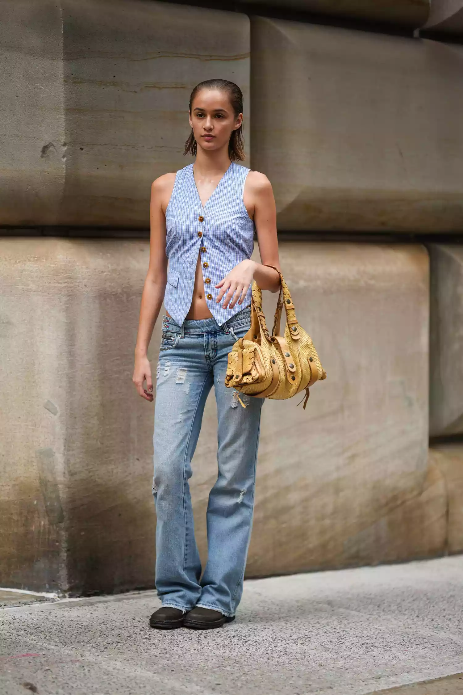 A model wears a blue waistcoat, a yellow bag, blue flared ripped jeans, brown shoes, a shopping bag